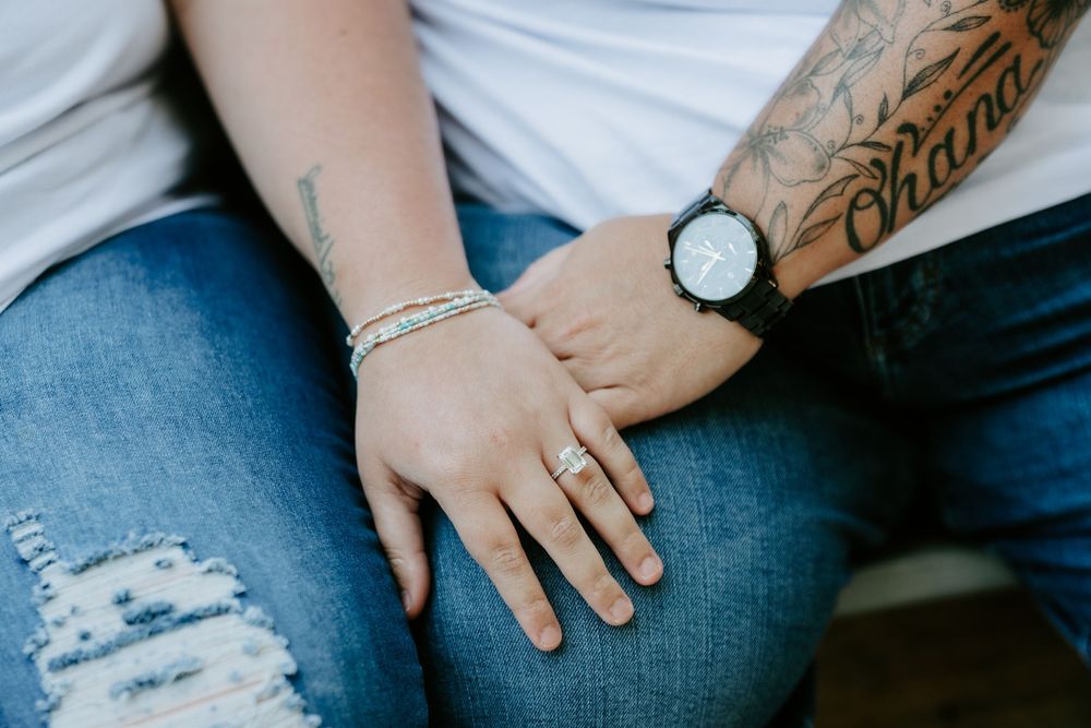 Close-up of two people in ripped jeans holding hands, one wearing a wristwatch and a tattooed forearm, the other with bracelets and a ring.