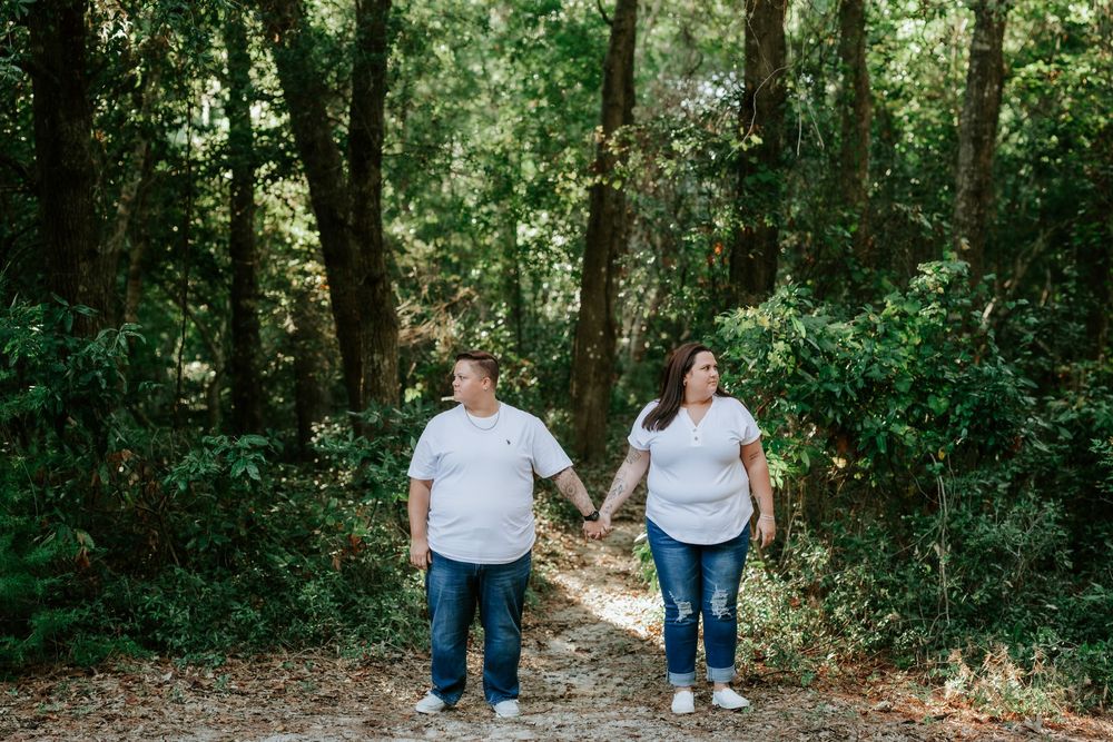 Couple in white shirts and jeans holding hands while walking along a sandy path through a green wooded forest.