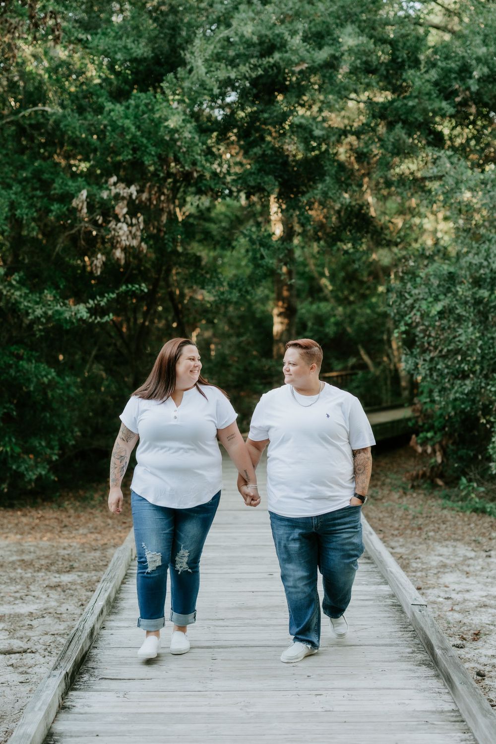 Couple in white polos and jeans hold hands while walking along a wooden boardwalk surrounded by green trees.