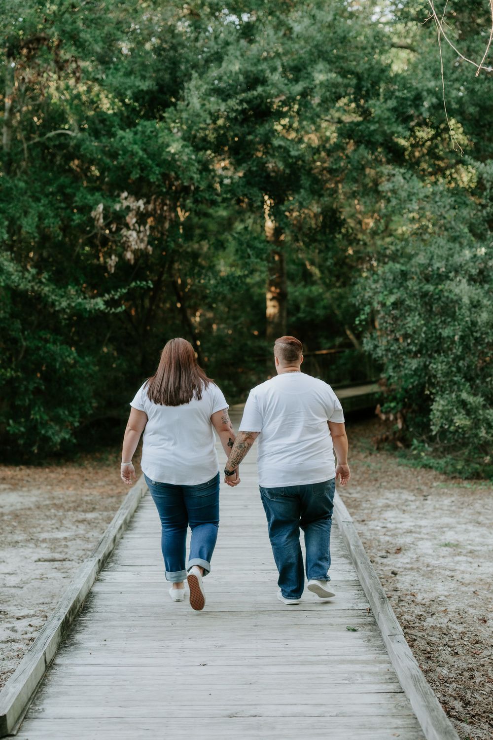 Two people in white shirts and jeans walk hand in hand away from the camera along a wooden boardwalk through a sunlit, tree-lined forest.