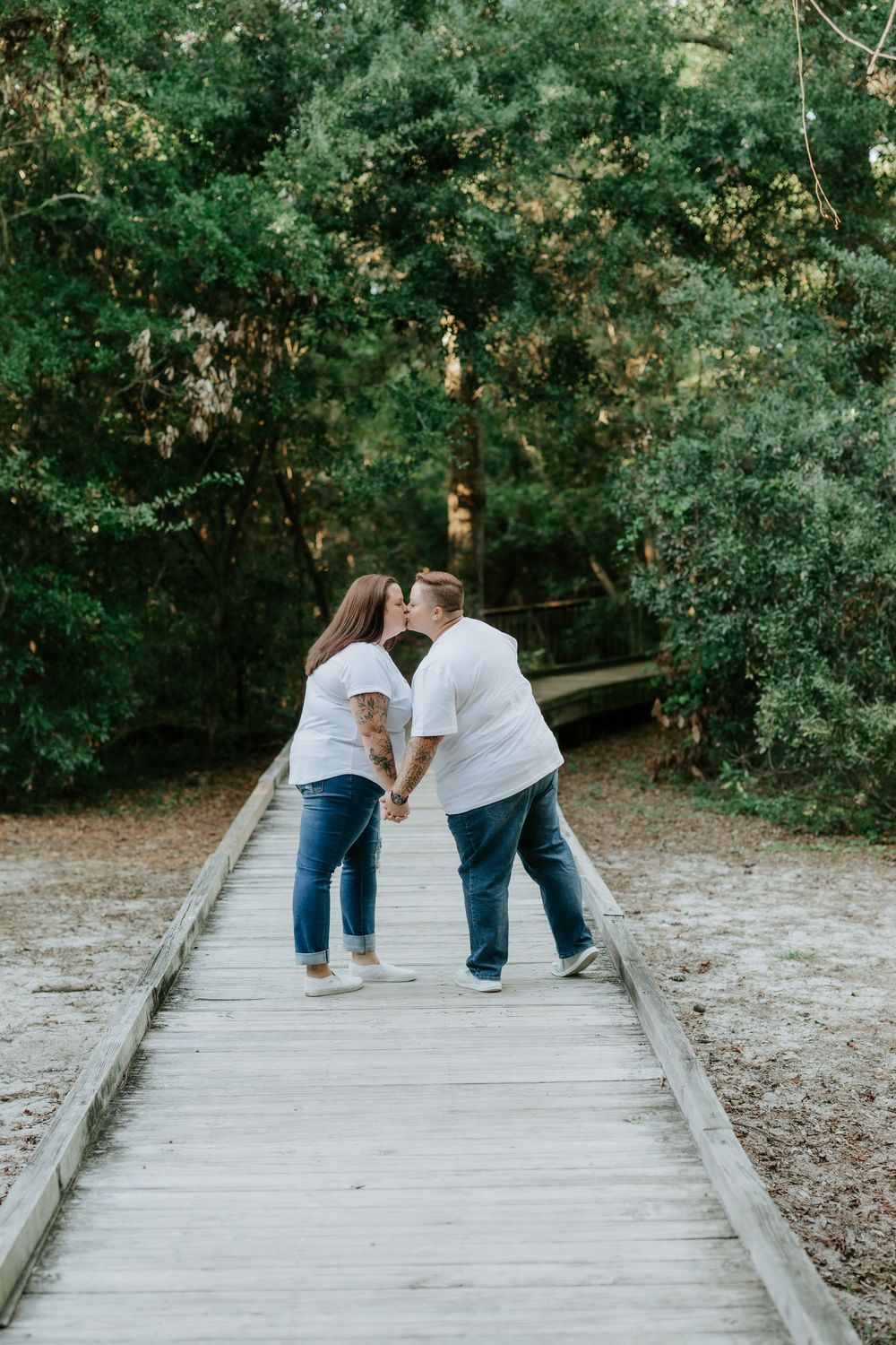 Couple in white shirts and jeans share a kiss while holding hands on a wooden boardwalk surrounded by green trees.
