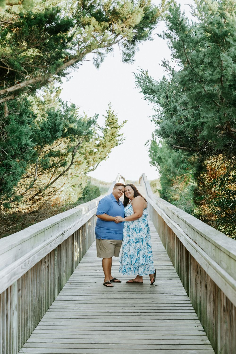 Smiling couple holding hands on a wooden boardwalk surrounded by coastal pine trees during an outdoor engagement photo session.