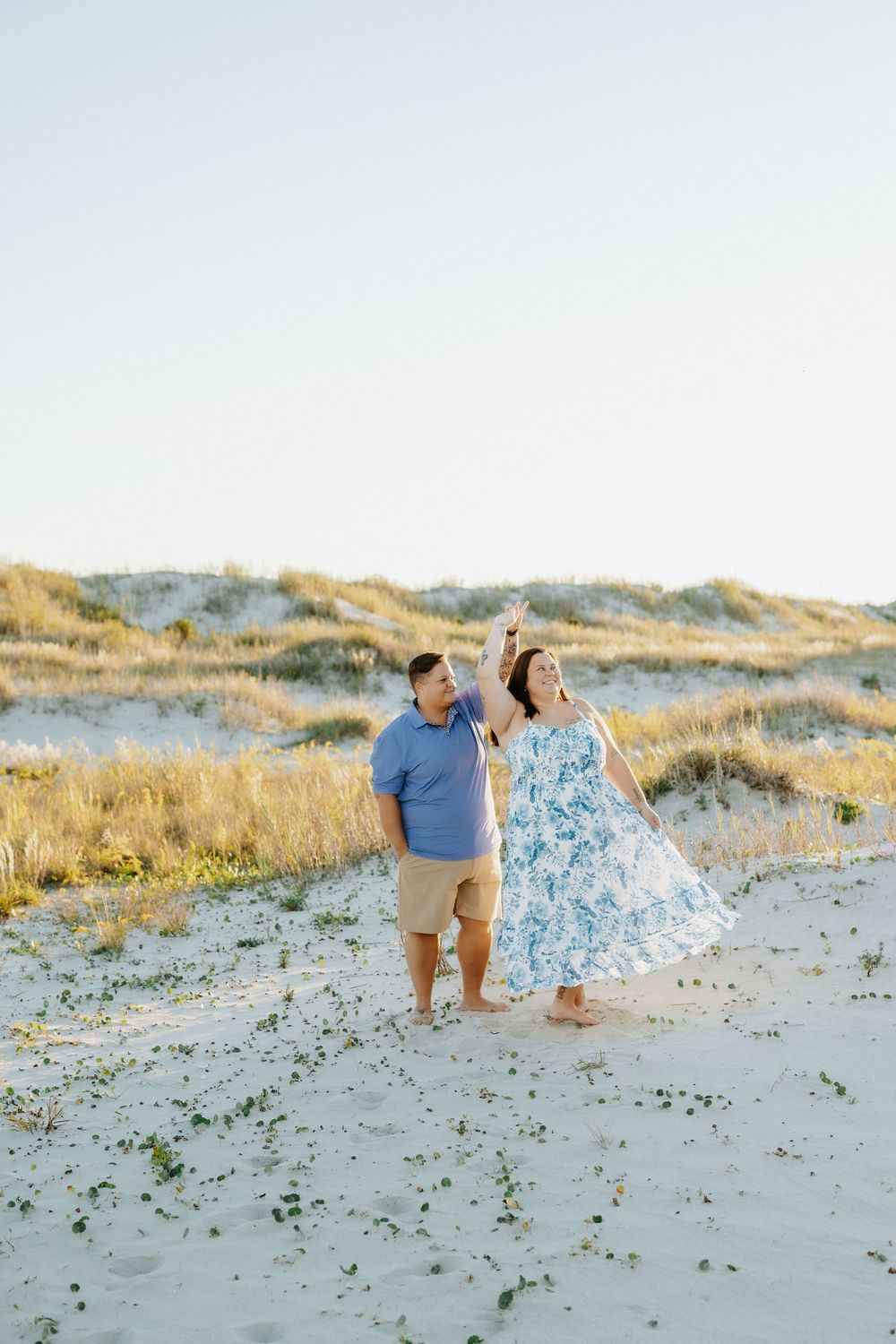 Couple twirls and dances barefoot on a sandy beach with dunes and sea grass during an engagement photo session at sunset.
