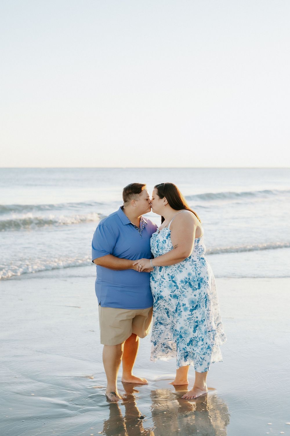 Couple in blue outfits share a kiss while holding hands barefoot in the shallow surf on a beach at sunset.
