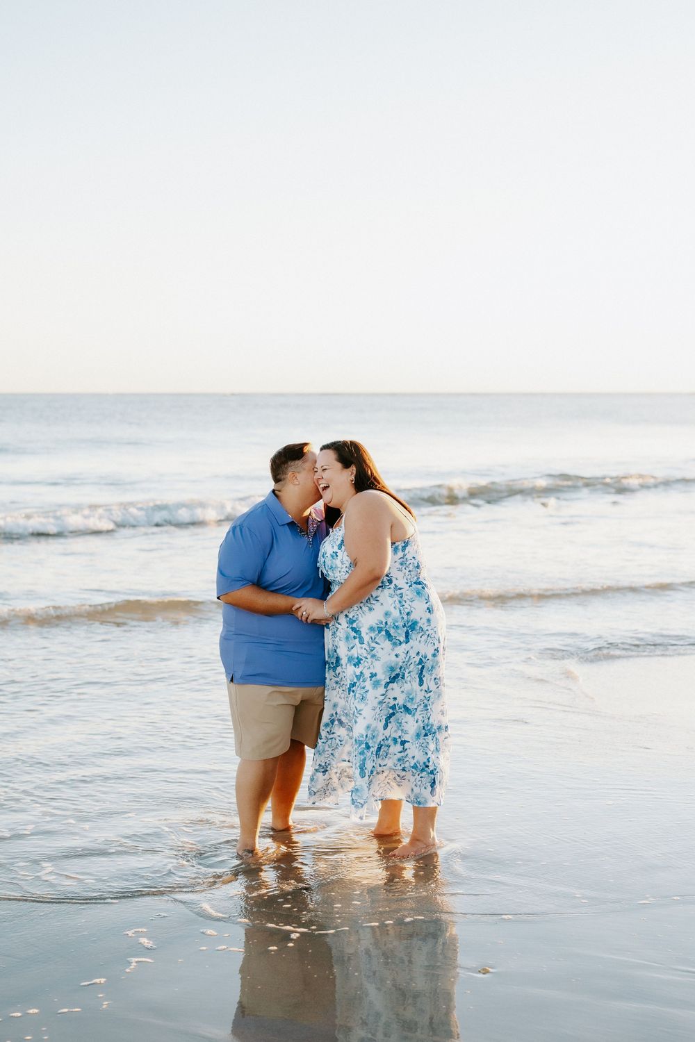 Couple in blue outfits embracing and sharing a kiss while standing barefoot in shallow ocean water on a calm beach at sunset.