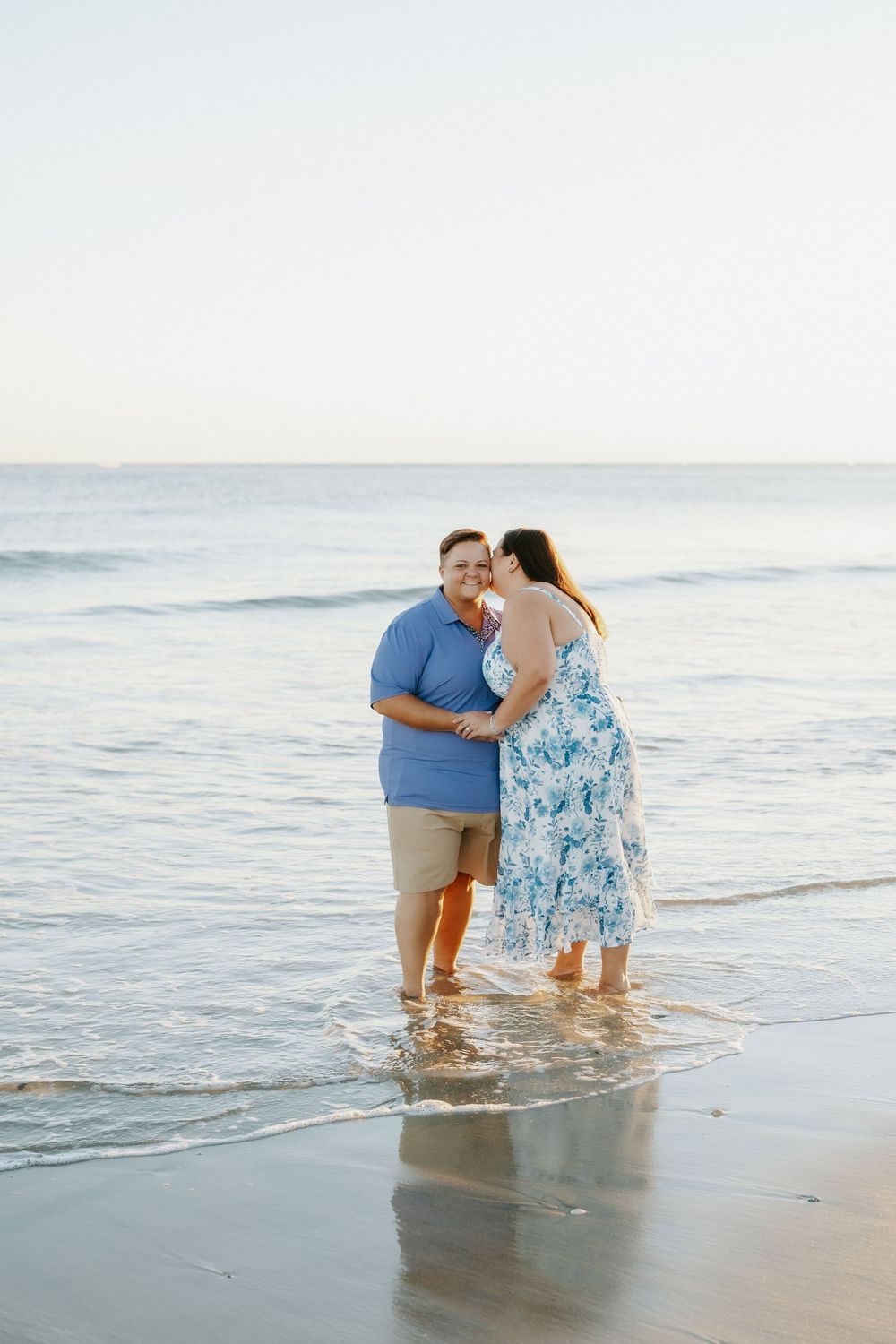 Couple in blue and floral outfits share a kiss while standing barefoot in shallow ocean water on a calm beach at sunset.