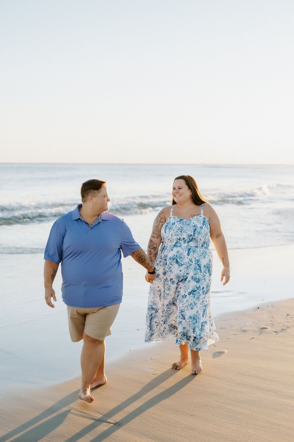 A smiling couple holds hands and walks barefoot along the sandy shoreline at sunset beside gentle ocean waves.