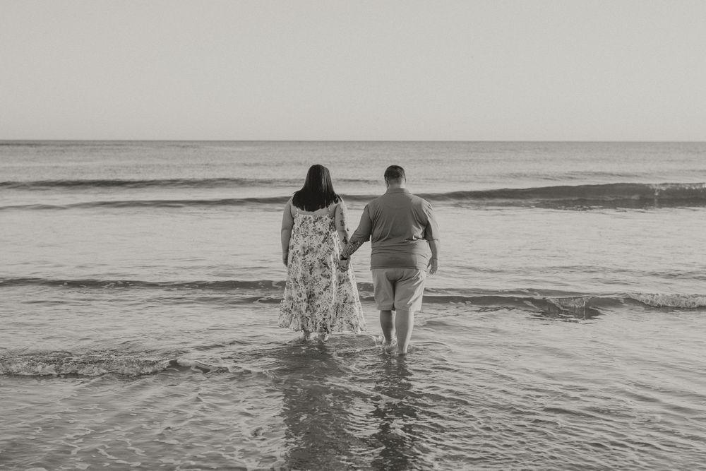 Couple holding hands as they wade into gentle ocean waves on a beach in a black and white engagement photo.