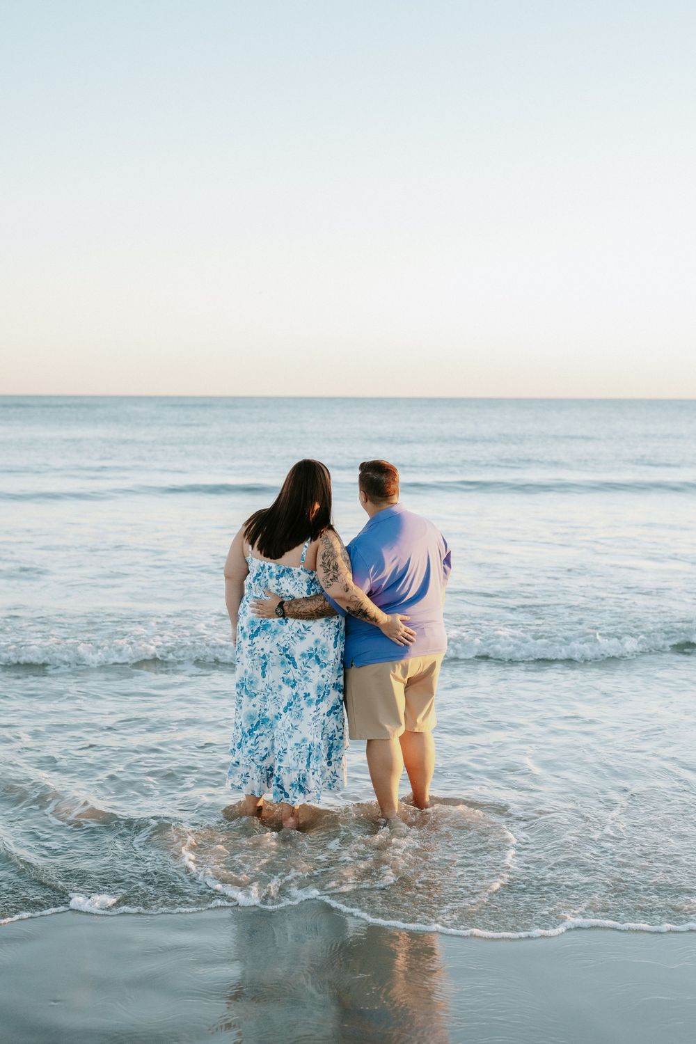 Couple embracing with arms around each other while standing barefoot in shallow ocean waves at sunset on a calm beach.