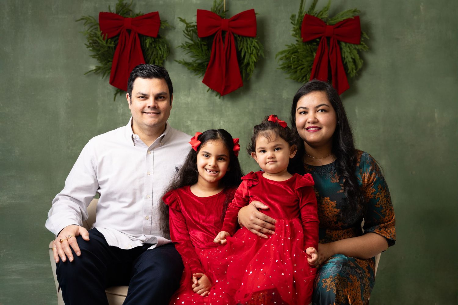 Family of four poses for a festive Christmas portrait with holiday greenery and red bows backdrop.
