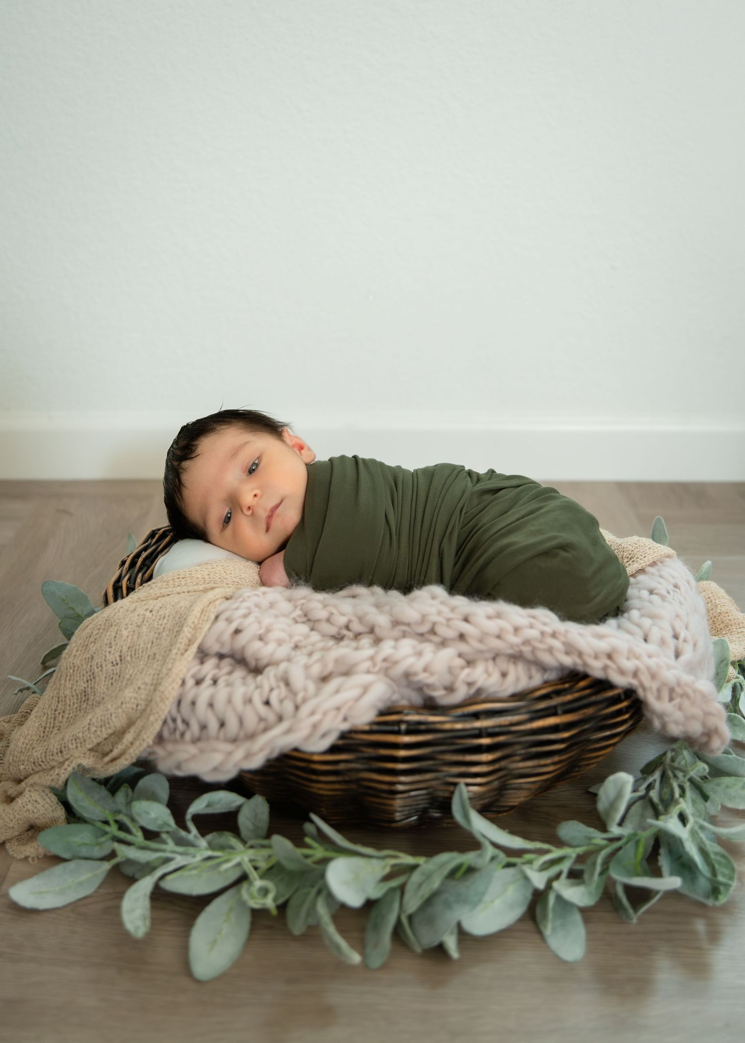 Newborn baby wrapped in olive green swaddle resting in a wicker basket with knit blanket and eucalyptus leaves.