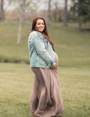 Smiling pregnant woman in denim jacket and flowing maxi dress poses outdoors in lush green park setting.