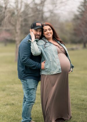 Smiling couple poses outdoors for maternity photo, woman in maxi dress and denim jacket showing baby bump.