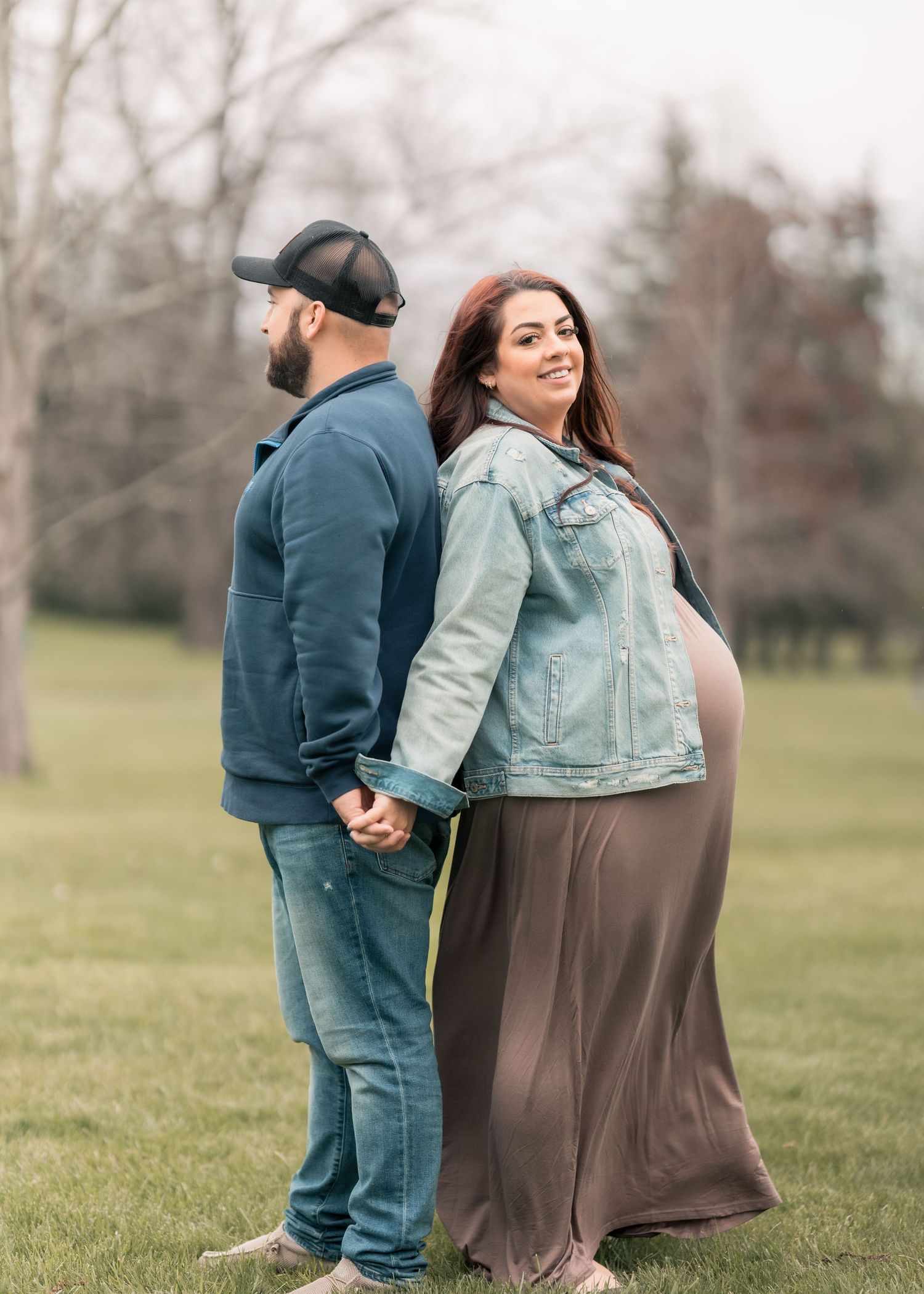 Pregnant woman in denim jacket and maxi skirt holds hands with man in blue hoodie and cap outdoors.