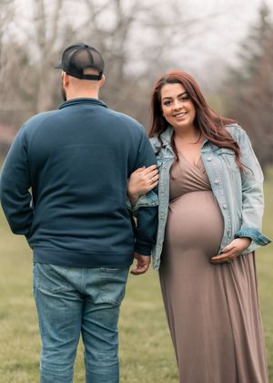Pregnant woman in maxi dress and denim jacket smiles at partner during outdoor maternity photo shoot.