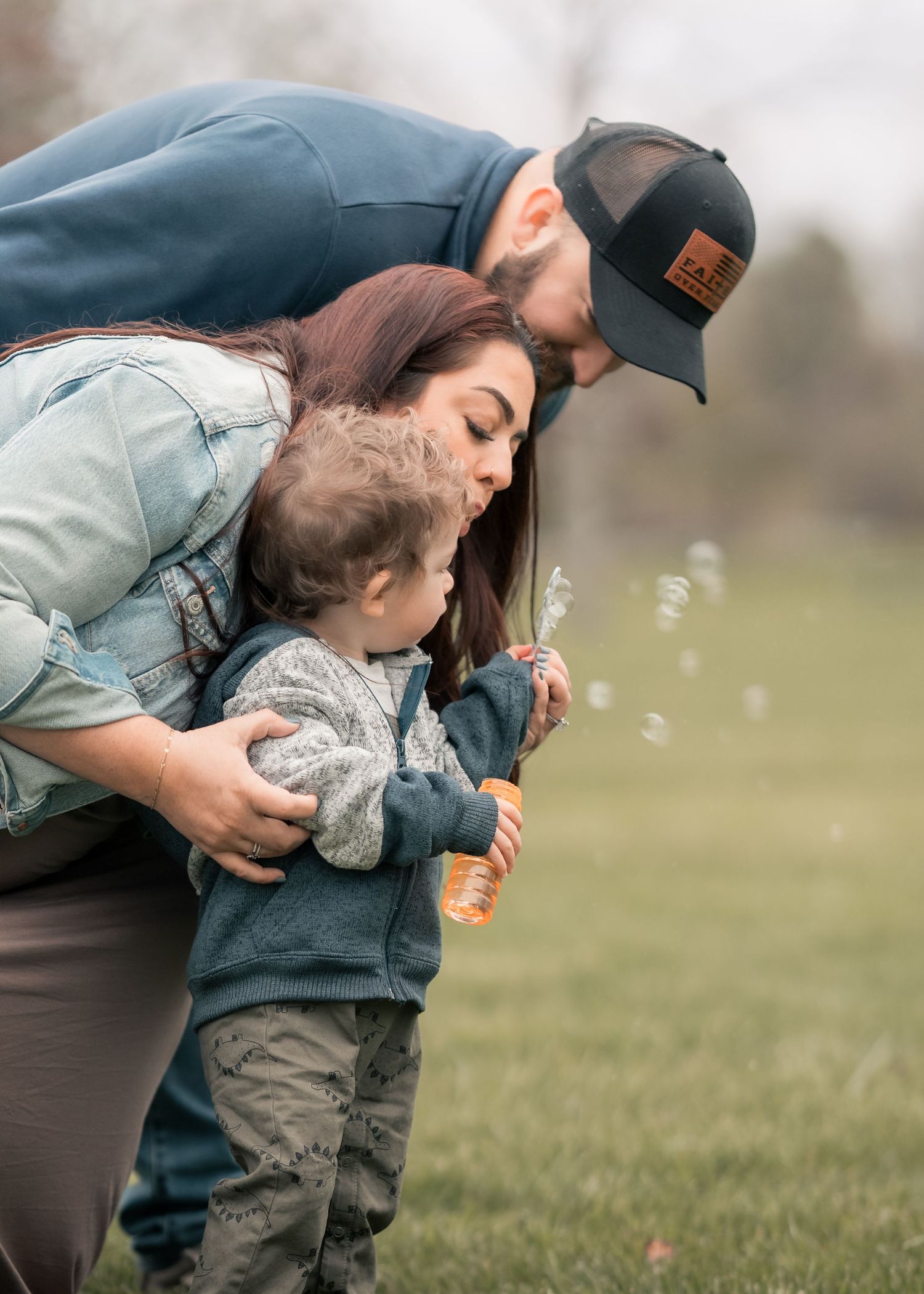 A family of three blowing bubbles together outdoors, with parents leaning over their toddler in a green park setting.