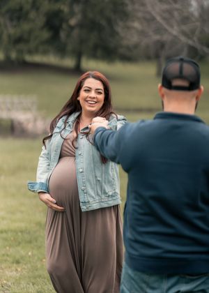 Pregnant woman in denim jacket and maxi dress smiles at partner during outdoor maternity photo session in green park.