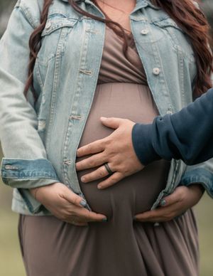 Pregnant woman in denim jacket and gray dress cradling baby bump with partner's hand gently resting on belly.