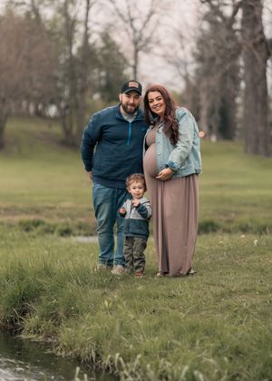 Pregnant woman in maxi dress and denim jacket poses outdoors with partner and toddler son for maternity family portrait.