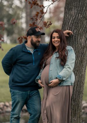 Couple posing outdoors by a tree, woman in flowy dress cradling baby bump, man in blue fleece and cap smiling at her.