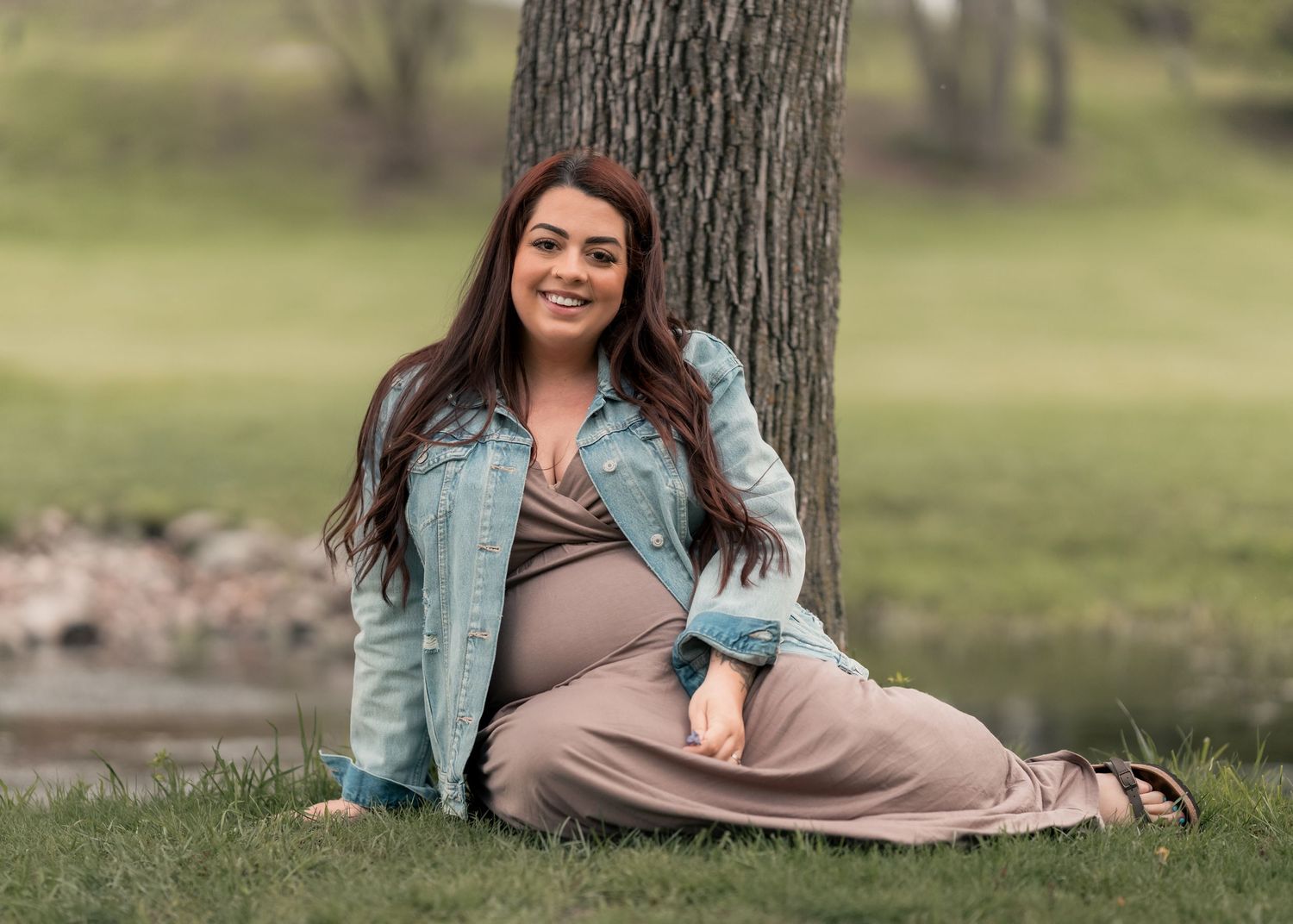 Pregnant woman in denim jacket and maxi dress sitting against tree in green park, smiling warmly.