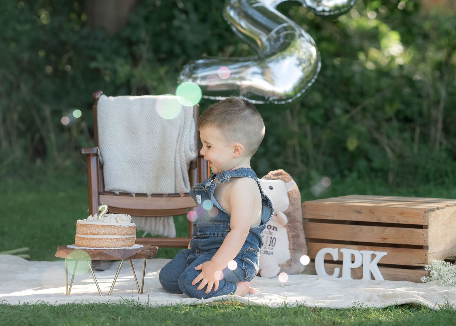 A baby boy in denim overalls sits on a blanket outdoors at a first birthday cake smash with CPK letters and balloons.