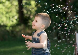 Toddler boy in denim overalls laughing and clapping hands while playing with colorful soap bubbles outdoors.