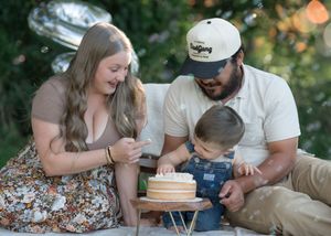 Family celebrating baby’s first birthday smash cake outdoors, parents smiling as toddler explores cake.