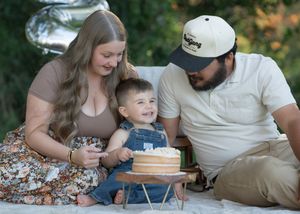 Family celebrating baby’s first birthday with a smash cake outdoors, parents smiling beside happy toddler.