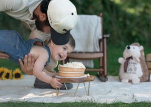 A baby smashes a birthday cake outdoors while an adult leans over to help, with sunflowers and a teddy bear nearby.