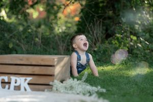 A joyful toddler boy laughing outdoors on green grass beside a wooden crate during a birthday photo session.