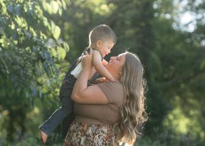 A mother lifts her young son outdoors, sharing a joyful nose-to-nose moment surrounded by lush green trees.