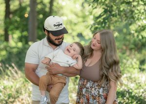 A happy young family with a man in a cap, a woman with long hair, and a laughing baby outdoors in a green park.