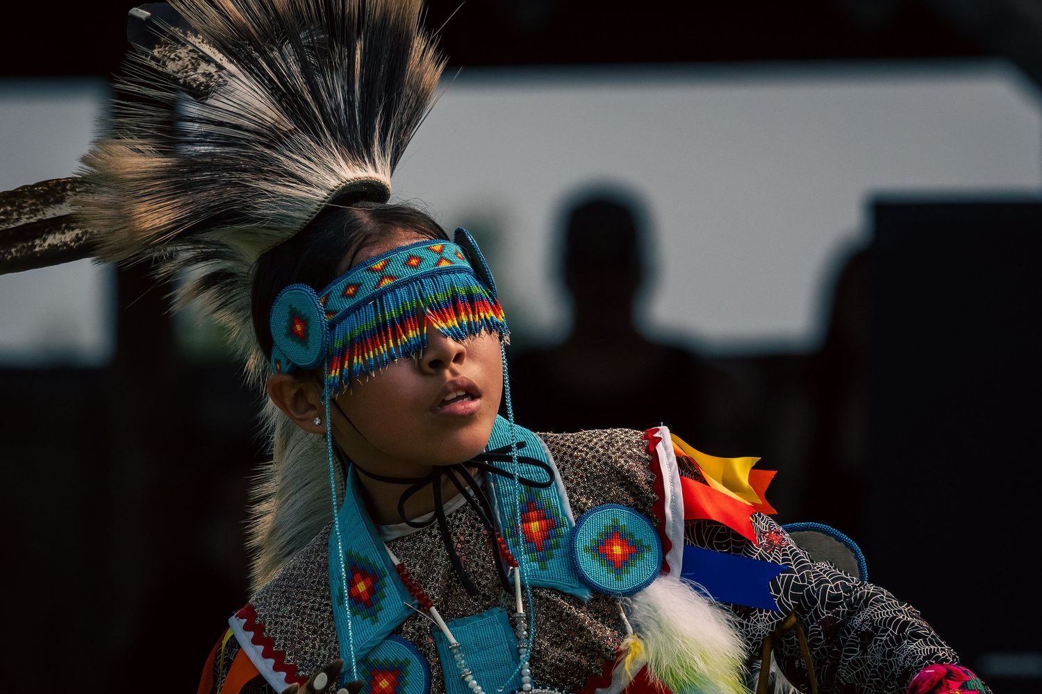 Dancer in vibrant turquoise beaded headband and feathered regalia performs at a powwow with silhouetted spectators in the background.