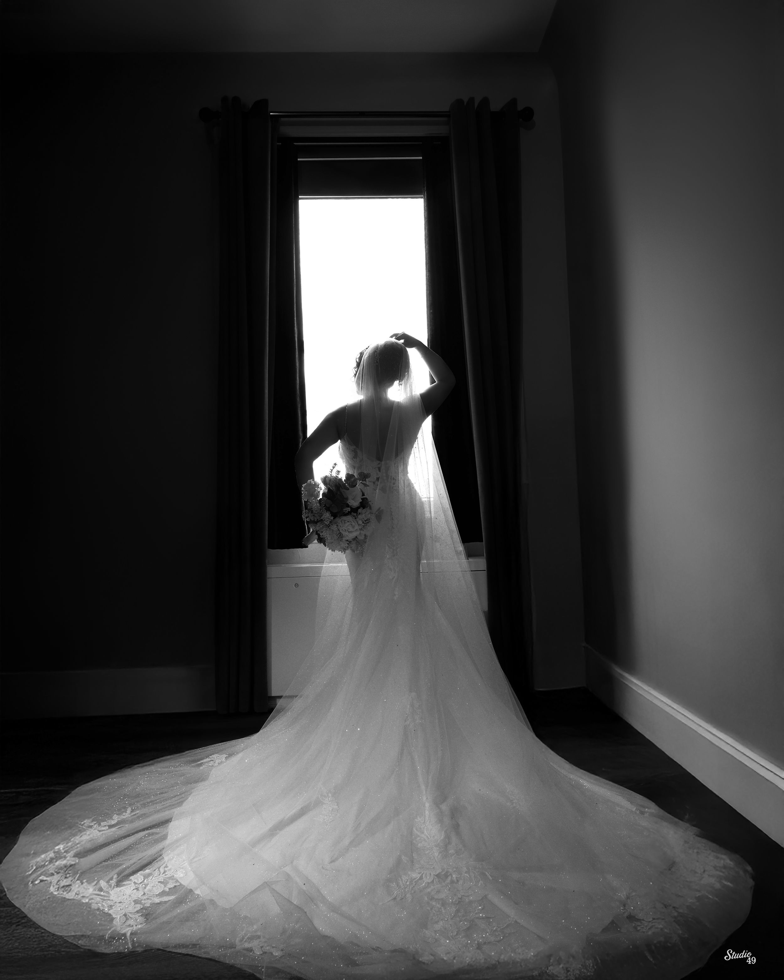 Black and white photo of a bride in a lace gown with a long train, holding a bouquet and standing by a sunlit window.