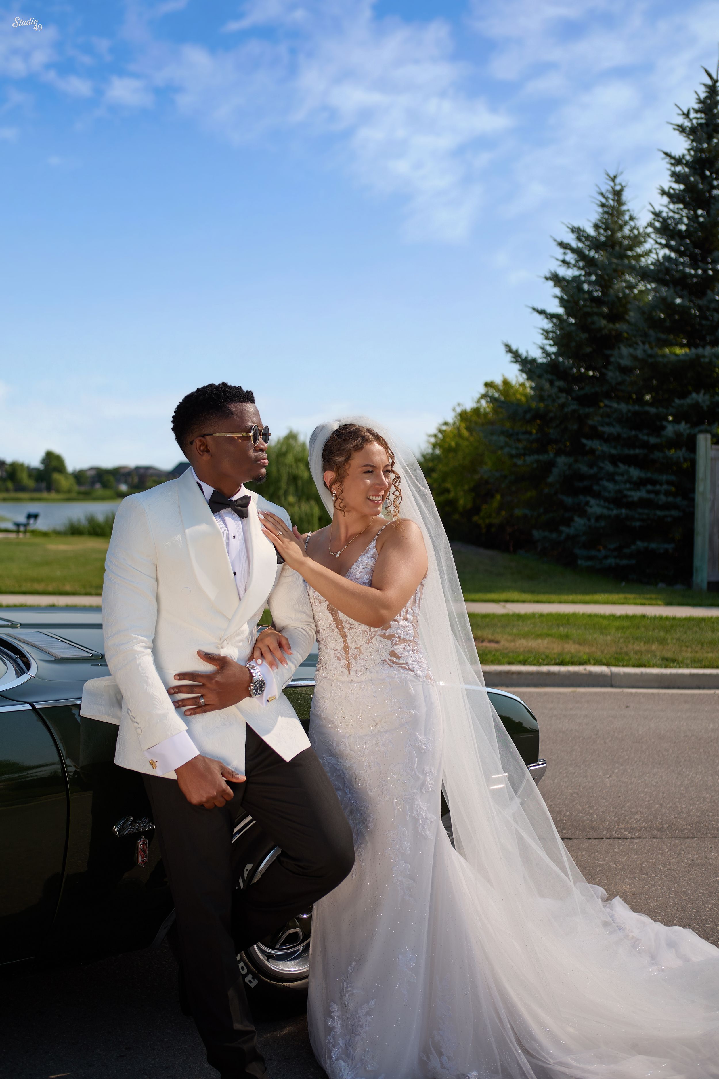 A smiling bride in a lace mermaid gown and veil poses with a groom in a white tuxedo jacket beside a vintage black convertible.