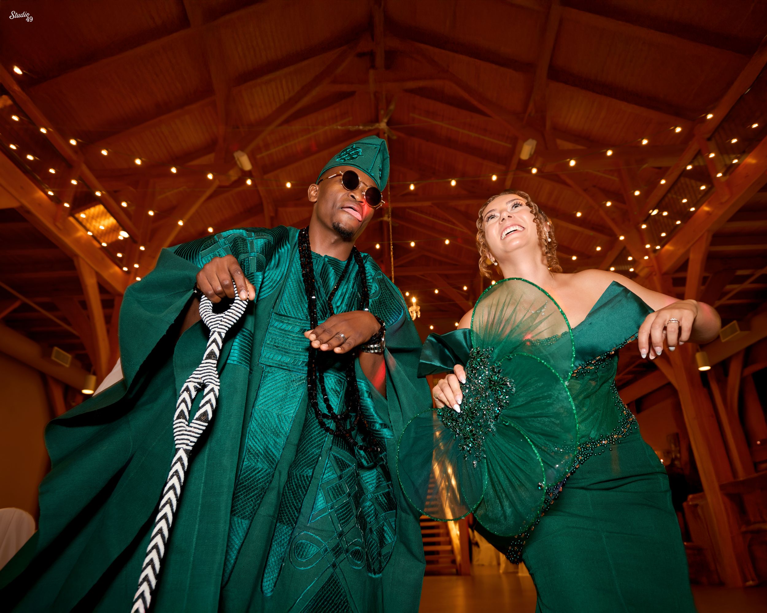 Smiling couple in matching emerald green traditional outfits dancing joyfully under a wooden beamed ceiling with twinkling string lights.