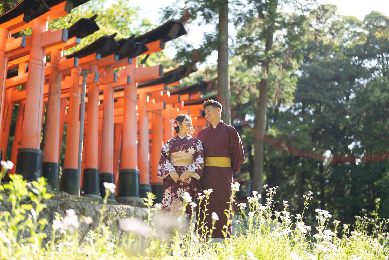 Couple in traditional Japanese kimono standing near vermilion torii gates surrounded by green trees and wildflowers at a Kyoto shrine.