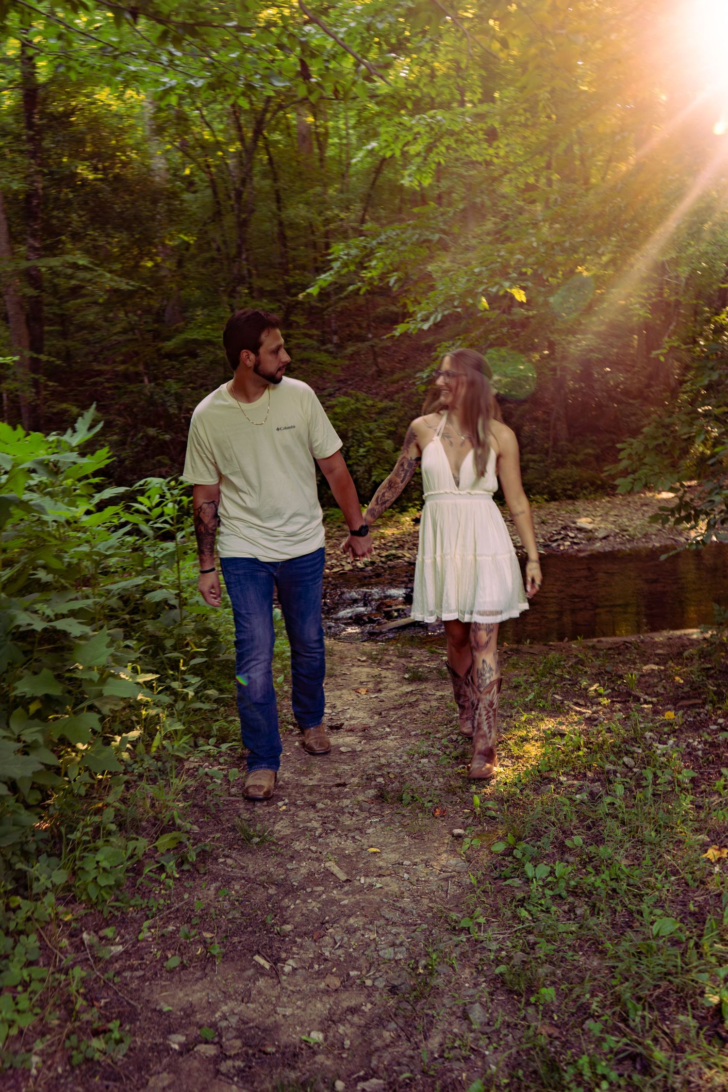 Couple holding hands and walking along a sunlit forest trail near a gentle creek.