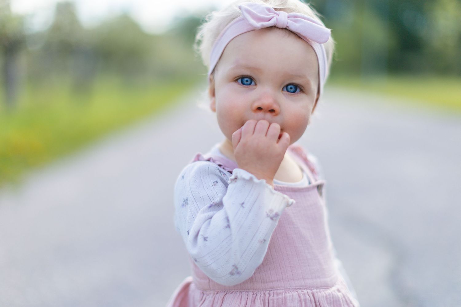 Adorable blue-eyed baby girl in pink dress and bow headband standing on a road, hand near mouth.