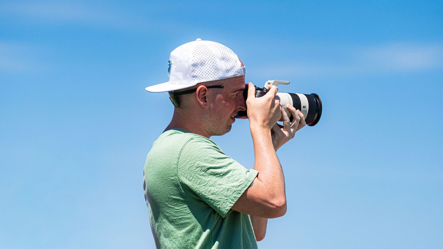 A photographer in a green tee and white cap holds a Sony camera with a long white lens to his eye, shooting against a clear blue sky