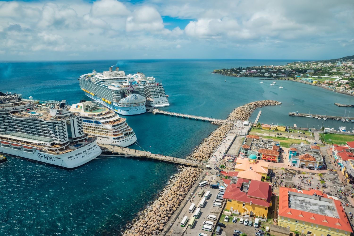 Aerial view of a tropical cruise port with multiple large ships docked along a pier surrounded by turquoise waters.
