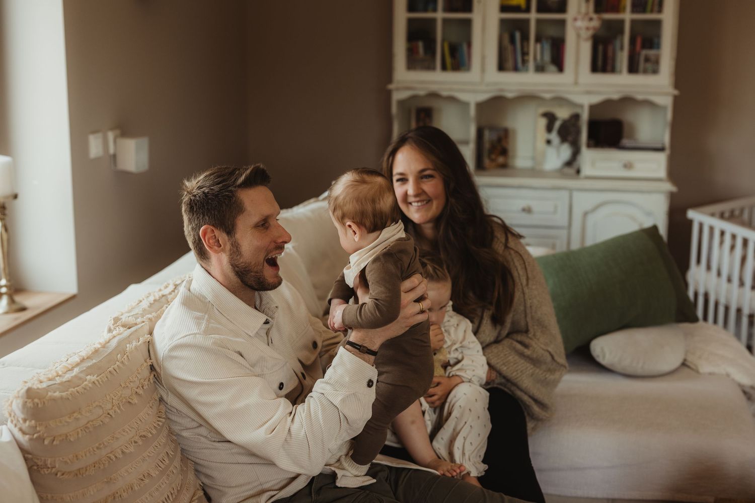 Happy young family with baby sitting on couch in cozy nursery room, sharing a joyful moment together.