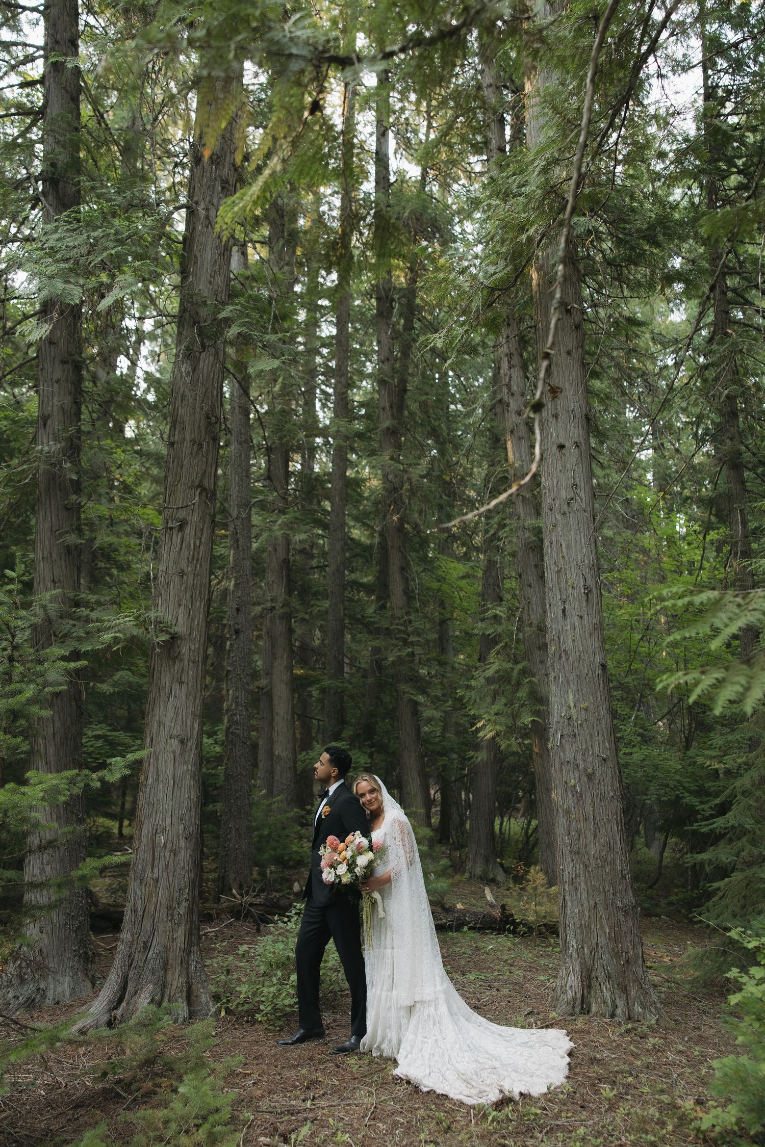 Bride and groom embracing among tall evergreen trees in a lush forest, bride holding a bouquet in a flowing white gown.