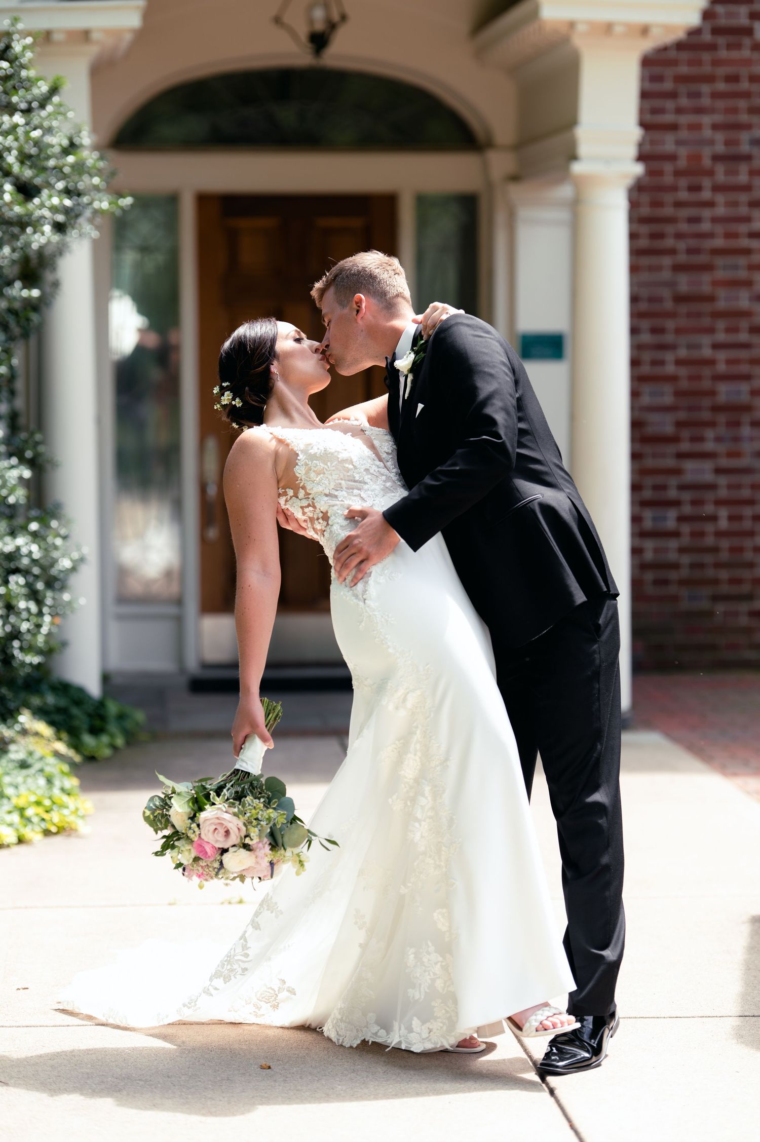 Bride and groom share a romantic kiss outside a brick building, bride holding a pink and white bouquet in a lace wedding gown.