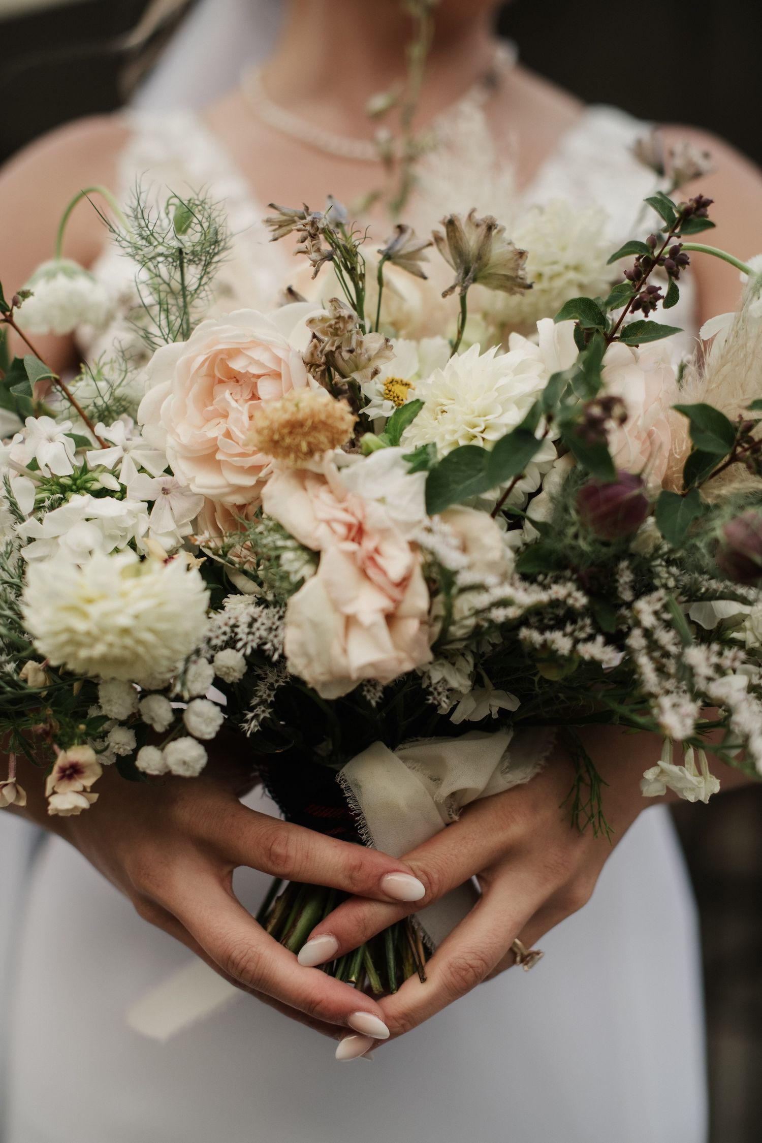 Bride holding a lush wedding bouquet with blush roses, white dahlias, and greenery, wearing a white gown and gold ring.