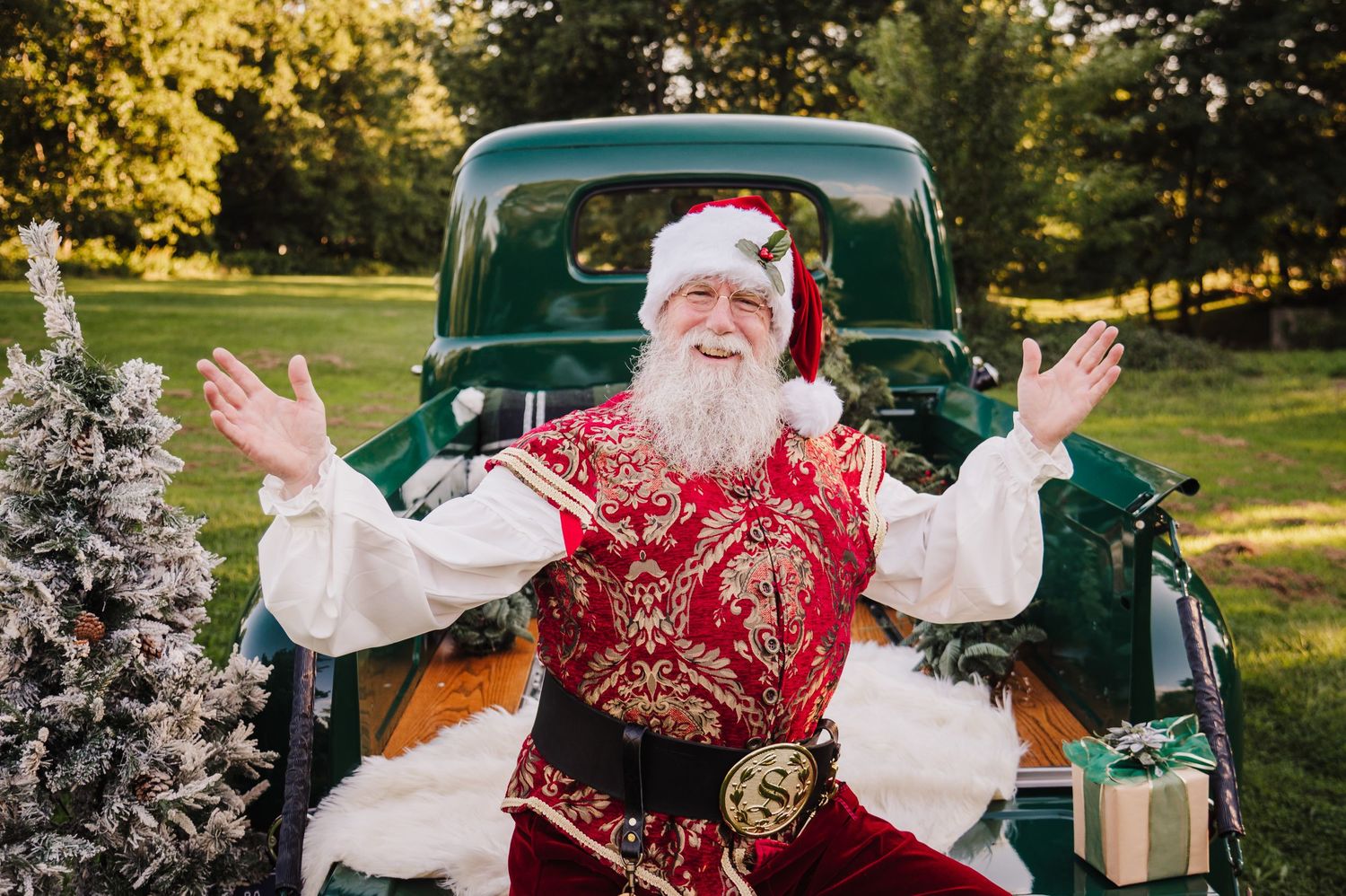 Santa Claus in ornate red suit poses with arms raised beside a vintage green truck outdoors.