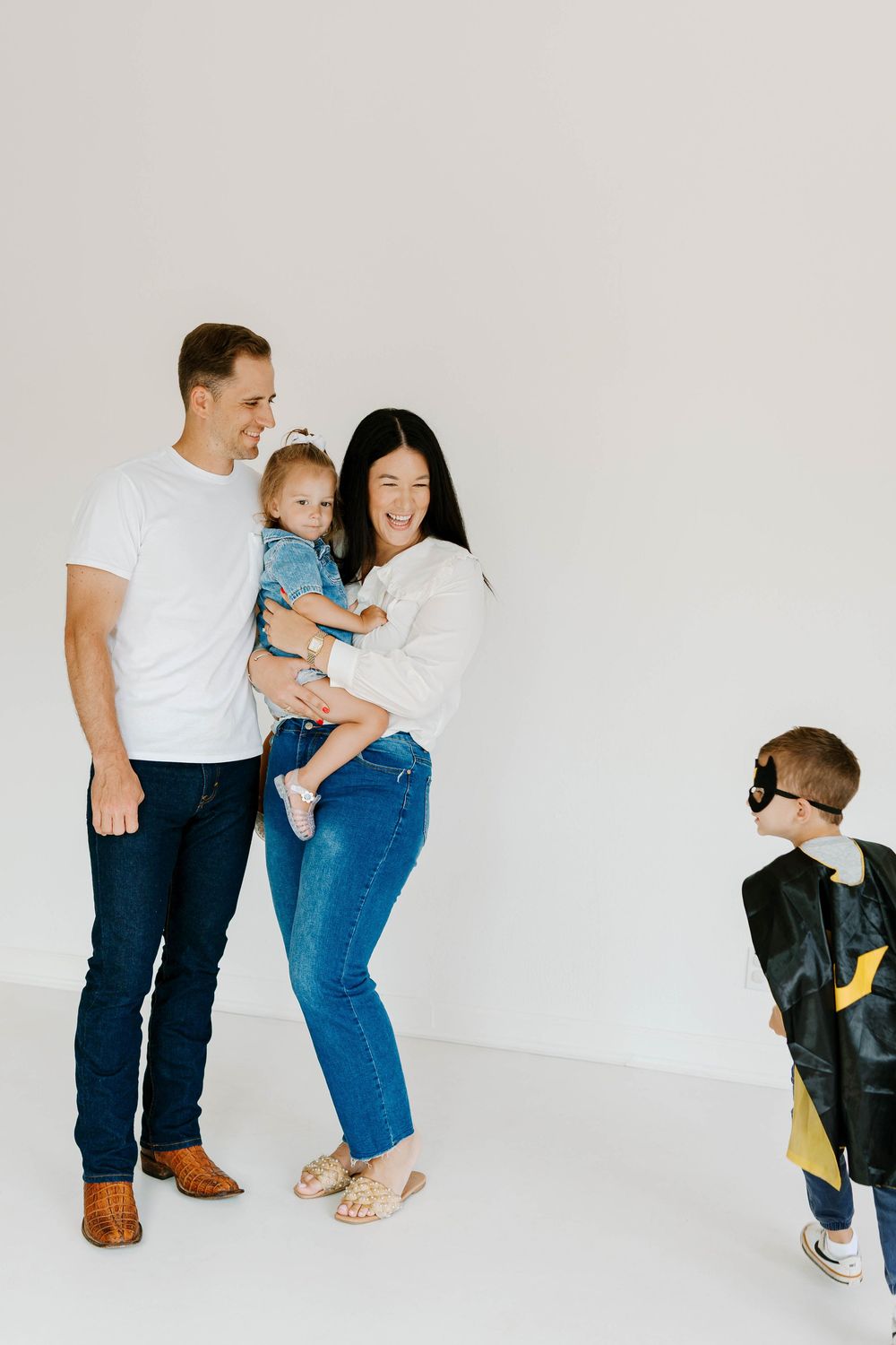 Family of four in casual white and denim outfits, toddler boy dressed as superhero in bright studio.