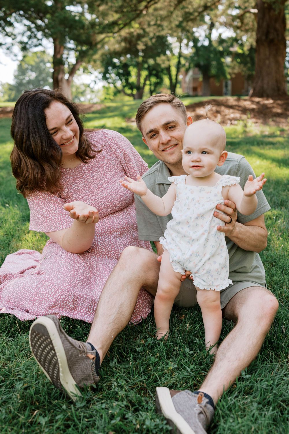 Smiling young parents sitting on grass in park with happy baby in white romper.