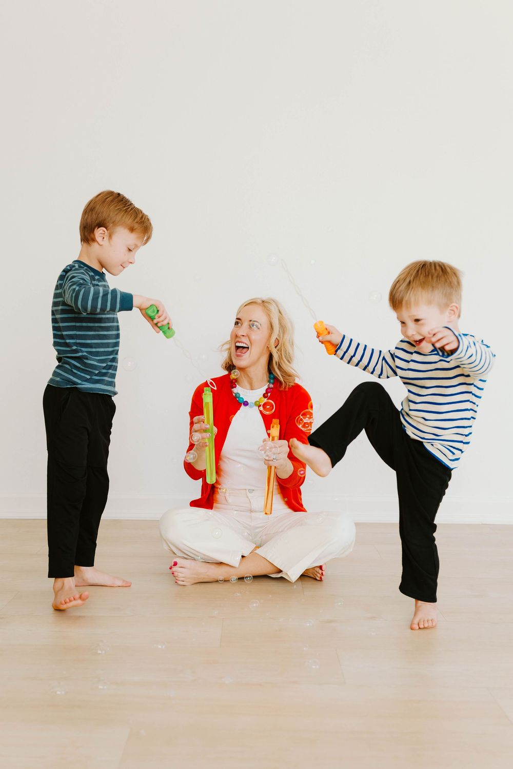 Laughing mother sitting cross-legged while two energetic boys play with toy weapons around her.